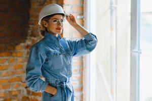 attraktiv kvinna konstruktion arbetstagare i hjälm. självsäker ung specialist i rutig blå skjorta i jeans stående i tömma rum. interiör design och renovering service foto