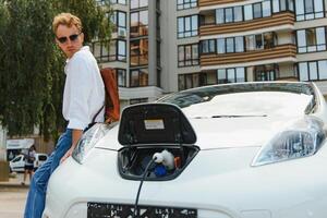 man laddning hans lyx elektrisk bil på utomhus- station i främre av modern ny stad byggnader foto