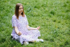 gravid kvinna håller på med yoga i de parkera. Sammanträde på de gräs. foto