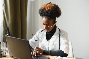 leende afrikansk amerikan kvinna läkare gp bär vit medicinsk täcka använder sig av bärbar dator dator på arbetsplats ger avlägsen uppkopplad samråd, arbetssätt på pc, hört patient i internet telemedicin chatt foto