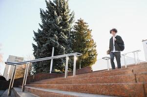 universitet.ler ung studerande man innehav en bok och en väska på en universitet bakgrund .ung leende studerande utomhus liv stil.stadsstudent. foto