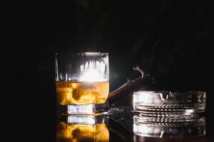 en glas av whisky eller bourbon med is kuber och en cigarr på en svart skiffer med rök foto