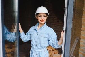 attraktiv kvinna konstruktion arbetstagare i hjälm. självsäker ung specialist i rutig blå skjorta i jeans stående i tömma rum. interiör design och renovering service foto