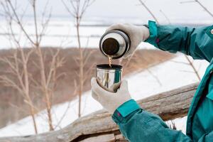 termos te kokande vatten Häll i en råna i vinter- till sola sig i natur, dryck en varm dryck utanför, de begrepp av en termos i de händer av en person till håll, Häll i vatten in i en glas. foto