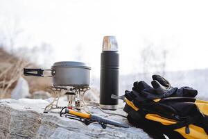 camping redskap stående på en sten, ryggsäck för vandring, värma handskar, termos varm te, kniv bushcraft, pott mat, gas brännare brand, camping Utrustning foto