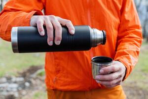 en turist man verkar till vara innehav en termos med varm te i hans händer, en man häller kaffe in i en råna i natur, turist Utrustning, vandring Utrustning, en Vakuum flaska. foto