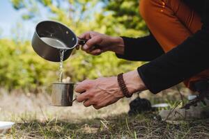 Häll i de vatten från de pott in i de glas. en turist i de skog häller kokande vatten från en pott in i en mugg, brygga te i natur, koka upp vatten, turist maträtter, Utrustning. foto