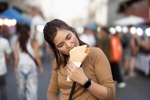 Lycklig ung asiatisk resande foodie kvinna njut av äter crepes på utomhus- natt marknadsföra gata mat Säljare foto