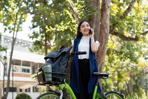 asiatisk affärskvinna med cykel använder sig av smartphone och Sammanträde utanför de kontor byggnad. kvinna pendling på cykel gå till arbete. eco vänlig fordon, hållbar livsstil begrepp foto