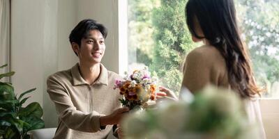 romantisk ung asiatisk par fattande med innehav blommor och leende i levande rum på Hem. falla i kärlek. valentine begrepp foto