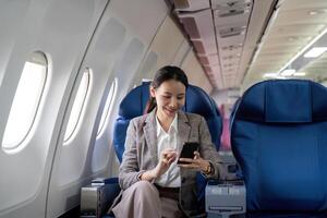 asiatisk ung affärskvinna framgångsrik eller kvinna entreprenör i formell kostym i en plan sitta i en företag klass sittplats och användningar en smartphone under flyg. reser och företag begrepp foto