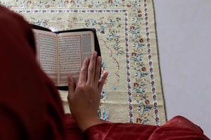 en muslim kvinna Sammanträde på en bön matta och läsning de quran med indonesiska översättning foto