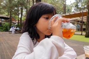 ett asiatisk liten flicka dricka en plast kopp av is te med natur eller träd bakgrund foto