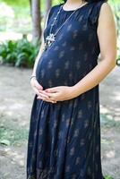 en gravid indisk lady poser för utomhus- graviditet skjuta och händer på mage, indisk gravid kvinna sätter henne hand på henne mage med en moderskap klänning på samhälle parkera, gravid utanför moderskap skjuta foto