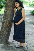 en gravid indisk lady poser för utomhus- graviditet skjuta och händer på mage, indisk gravid kvinna sätter henne hand på henne mage med en moderskap klänning på samhälle parkera, gravid utanför moderskap skjuta foto