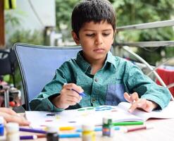 smart indisk liten pojke prestera tumme målning med annorlunda färgrik vatten Färg utrustning under de sommar semester, söt indisk unge håller på med färgrik tumme målning teckning på trä- tabell foto