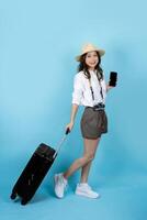 skön ung asiatisk turist kvinna gående med vagn resväska och smartphone, studio skott blå Färg isolerat bakgrund foto