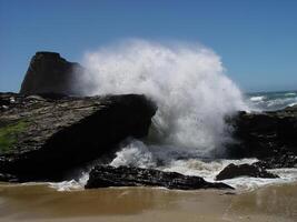 Vinka kraschar över stenar på sandig strand blå himmel foto