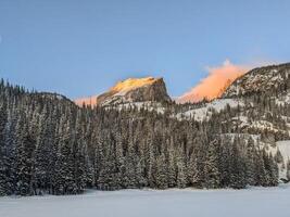 alpina morgon- glöd på en berg topp med en snöig sjö och vintergröna träd foto