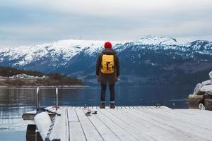 resenär man med en gul ryggsäck bär en röd hatt som står på bakgrunden av berg och sjö träbrygga. resa livsstilskoncept. skjuta från baksidan foto