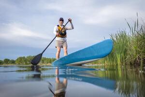 senior manlig paddlare är paddling en stå upp paddelbräda på en lugna sjö i vår, groda perspektiv från ett verkan kamera på vatten nivå foto