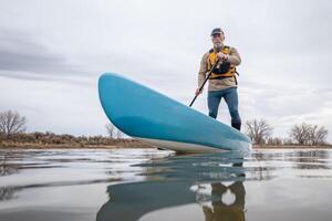 solo- senior manlig paddling en stå upp paddelbräda på en sjö i tidigt vår, groda perspektiv från ett verkan kamera på vatten nivå foto