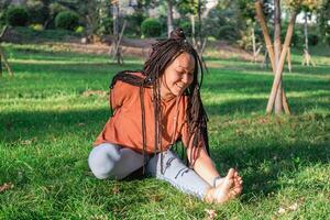 skön ung kvinna med lång afrikansk flätor är håller på med yoga utanför i en parkera. begrepp av friska livsstil. foto