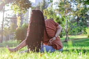 skön ung kvinna med lång afrikansk flätor är håller på med yoga utanför i en parkera. begrepp av friska livsstil. foto