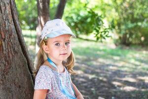 liten flicka i en blommig klänning och hatt lutande mot en träd i de parkera foto