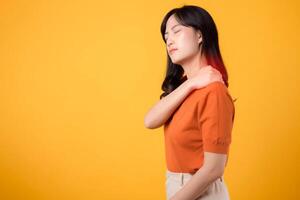 vaksam ung asiatisk kvinna i henne 30-talet, bär ett orange skjorta, innehar henne smärta axel på gul bakgrund. nacke värk terapi medicinsk kontor syndrom begrepp. foto