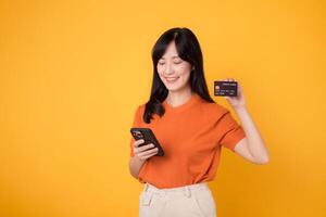 dynamisk ung asiatisk kvinna i henne 30-talet, använder sig av smartphone och innehav kreditera kort på gul bakgrund. effektiv uppkopplad handla. foto