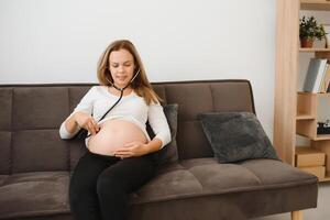 gravid kvinna använder sig av stetoskop granskning henne mage på soffa, Lycklig gravid kvinna begrepp. foto