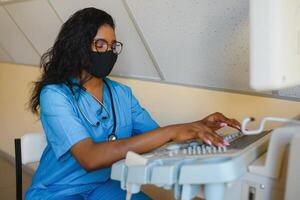 ung glad afrikansk kvinna operatör i mask av ett ultraljud läser in maskin analyserar diagnostik resultat av patient. ung leende afrikansk läkare arbetssätt på en modern ultraljud Utrustning. foto