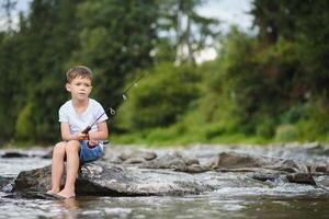 söt pojke i vit t skjorta fiske i de flod och har roligt, ler. semester med ungar, högtider, aktiva helger begrepp foto