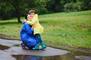 mor och barn, pojke, spelar i de regn, bär stövlar och regnrockar foto