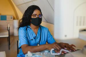 ung kvinna afrikan-amerikan läkare arbetssätt på modern ultraljud Utrustning. operatör av ultraljud läser in maskin Sammanträde och ser på de övervaka, väntar för patient. foto