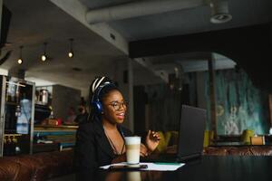 mänsklig och teknologi. ung afrikansk kvinna skriver på bärbar dator, textning vänner via social nätverk. studerande flicka bläddring internet, använder sig av fri wi-fi, Sammanträde på Kafé nära fönster på solig dag foto