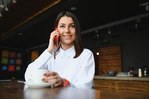 ung charmig kvinna kallelse med cell telefon medan Sammanträde ensam i kaffe affär under fri tid, attraktiv kvinna med söt leende har talande konversation med mobil telefon medan resten i Kafé foto