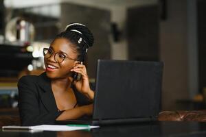 ung flicka i glasögon otroligt ser i bärbar dator på Kafé. afrikansk amerikan flicka Sammanträde i restaurang med bärbar dator och kopp på tabell. porträtt av överraskad lady med mörk lockigt hår i hörlurar foto
