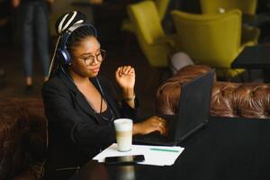 Lycklig svart flicka i trådlös hörlurar studerar uppkopplad, använder sig av bärbar dator och tar anteckningar, Kafé interiör, kopia Plats foto