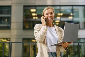Lycklig kvinna chef talande med klient och arbete på bärbar dator på modern kontor byggnad bakgrund foto