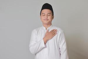 porträtt av religiös asiatisk man i koko skjorta eller vit muslim skjorta och svart keps, placering hand på hjärta, känsla mycket tacksam. isolerat bild på grå bakgrund foto