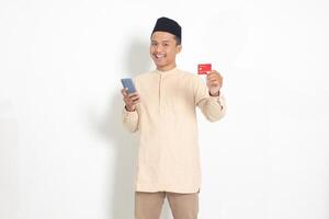 porträtt av attraktiv asiatisk muslim man i koko skjorta med kalott innehav en mobil telefon och presenter kreditera kort. isolerat bild på vit bakgrund foto
