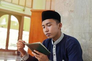 religiös asiatisk man i muslim skjorta och svart keps läsning de helig bok av quran i de offentlig moské foto
