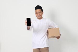 porträtt av Lycklig asiatisk muslim man i koko skjorta med peci bärande kartong låda medan som visar tom skärm mobil telefon för falsk upp. gående Hem för eid mubarak. isolerat bild på vit bakgrund foto