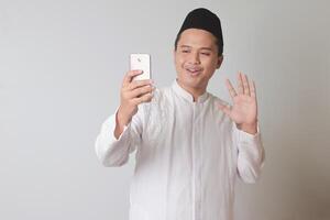 porträtt av asiatisk muslim man i vit koko skjorta med kalott tar bild av han själv eller selfie, ordspråk Hej och vinka hans hand. isolerat bild på grå bakgrund foto