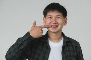 Lycklig asiatisk tonåring pojke pekande på tandställning. leende asiatisk tonåring man pekande på hans dental tandställning på en ljus grå bakgrund. porträtt stilig man i studio begrepp. foto