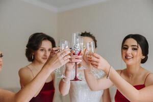 Foto med brudtärnor dricka champagne från glasögon i de morgon- i en skön hotell. Foto av en skön ung brud och henne vänner i matchande klänningar dricka champagne innan de bröllop.