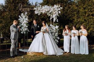 en bemästra av ceremonier ger en Tal medan en brud och brudgum håll händer nära en vit blommig båge foto