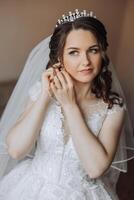 skön ung brud bär örhängen innan bröllop ceremoni på Hem. en brud i en vit bröllop klänning sätter på örhängen i henne rum i de morgon. foto